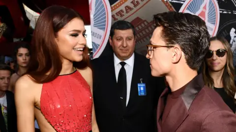 Getty Images Zendaya and Tom Holland promoting Spider-Man: Far From Home together in 2019. She is wearing a red dress and he has on a purple suit as people look on in the background