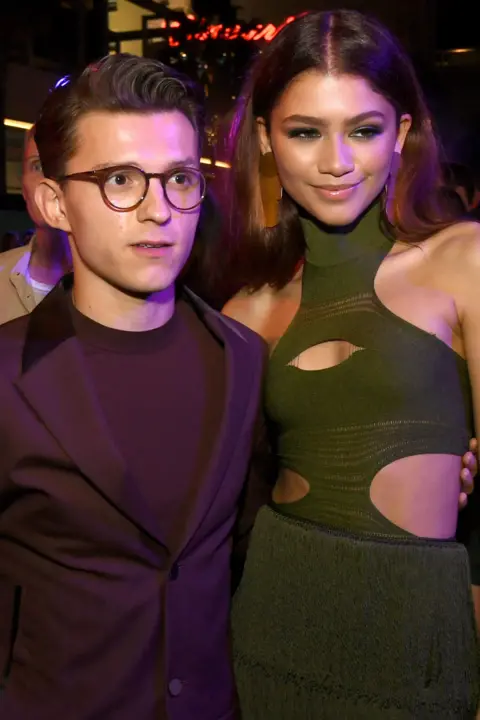 Getty Images Zendaya and Tom Holland promoting Spider-Man: Far From Home together in 2019. Holland is wearing glasses and has his arm around Zendaya's side. She is wearing a green turtle neck dress