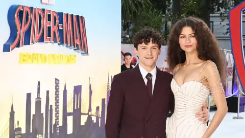 Getty Images Tom Holland and Zendaya promoting Spider-Man: Brand New Day at Wednesday's London premiere. He has a dark suit on while she is wearing a white dress. He has his arm around her waist and they are both stood in front of a film poster.