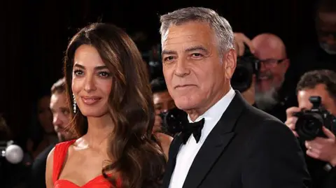 Getty Images Amal Clooney is dressed in a red dress while husband George can be seen in a tuxedo and black bow tie next to her. A bank of photographers are in the background out of focus