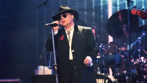 Getty Images Van Morrison is dressed in black and is wearing a black hat and shades while singing on stage 
