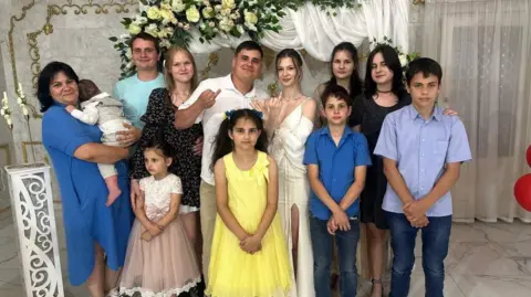 X/ Volodymyr Zelensky Twelve family members smile at the camera while what appears to be a bride and groom hold up their traditional wedding ring fingers