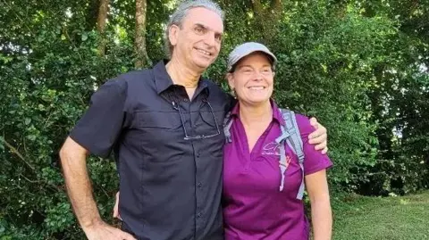 An image of Langlois and Marchand standing side by side with lush greenery behind them. He has grey hair and is wearing a black short-sleeved button up shirt with his arm around Dominique, who is wearing a cap, a backpack and a bright purple collard t-shirt. 