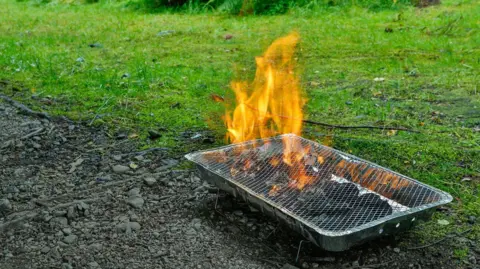 A disposable barbecue straddles a soily patch and grass as it burns.