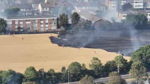 Aerial view of a grass fire in Romford showing a large area of blackened ground beside homes and apartment blocks. Smoke rises from the burn scar as firefighters work along the fire edge near residential properties.