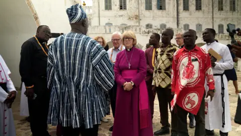 BBC Dame Sarah Mullally, a woman with short blond hair and a fringe, wearing reddish-purple robes and a gold cross necklace. She stands in front of sand-coloured buildings and is surrounded by half a dozen Ghanaian men.