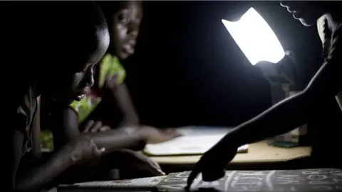 Three children study by the light of a bright lamp in a dark room, with one child pointing at a page while the others lean over their books 