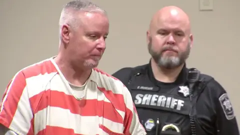 Colin Gray, in a white and orange stripped prison jumpsuit, with short grey cropped hair, is seen in profile. A sheriff's deputy in his black uniform is behind him 