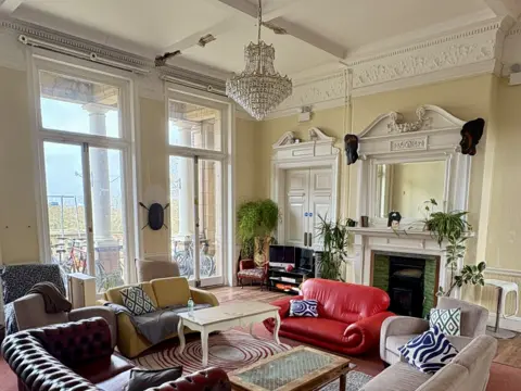 Katrina A large living room painted white with a chandelier hanging from the ceiling, a fireplace. 