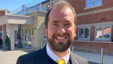 Tim Bearder has short brown hair and a brown beard. He is wearing a black blazer and jumper over a yellow tie and white shirt.