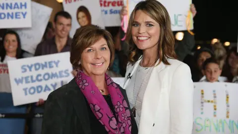 Getty Images Savannah, all in white, and Nancy Guthrie in a black suit jacket and pink patterned scarf, smile as they stand shoulder to shoulder. People behind them hold up signs. 