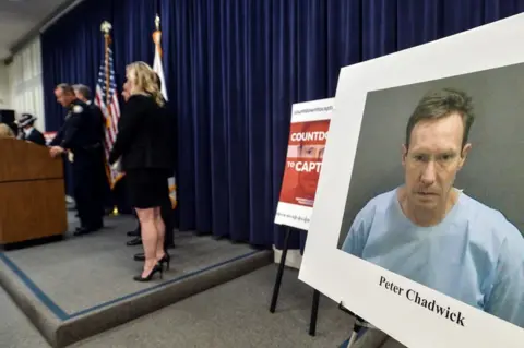 Getty Images A row of police officers are on stage addressing a room of journalists (out of view) and there is a picture mounted on a board with the caption Peter Chadwick typed underneath his mugshot, where he's wearing a pale blue top.