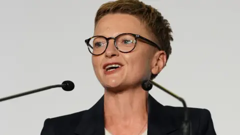 PA Media Bev Craig has short spiky red hair, dark glasses and wears a dark jacket with white roll-neck top.