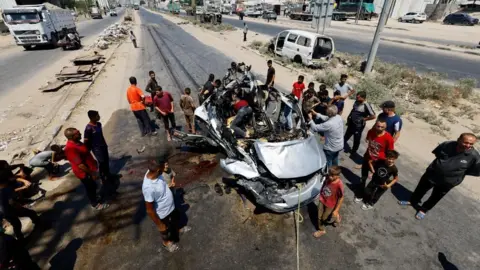 Reuters A large group of men surround a destroyed car
