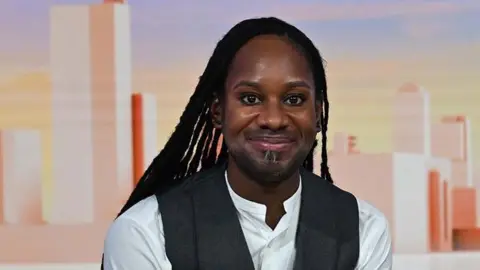 BBC Head shot showing Professor Jason Arday sitting in the Sunday with Laura Kuennsberg studio. He is smiling at the camera with long black locs, wearing a white shirt with a grey waist coat.