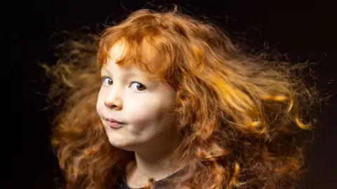 Kieran Dodds Close-up portrait of a young child with long, curly red hair flowing outward as if caught by a breeze. The child is turned slightly to one side and looking toward the camera, wearing a dark top against a dark, softly blurred background. Warm lighting highlights the texture and movement of the hair.