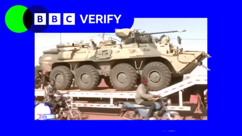 BBC A Russian military vehicle being transported through the Malian capaitl, Bamako. Men on motorcycles are riding alongside it. 