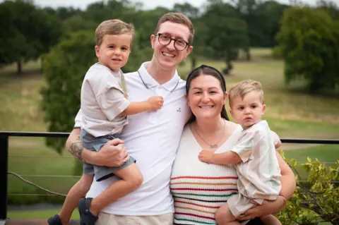 Alex West Four people standing together outdoors in the foreground, with two adults in the centre and a child held on each side. A man wears glasses and a white polo shirt, while a woman wears a sleeveless striped top. A metal railing runs behind the group, with grass, trees and open parkland visible in the background.
