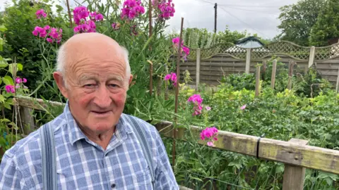 BBC Elderly man in blue check shirt looking into the camera. He's smiling and is standing in front of a garden with pink flowers. 