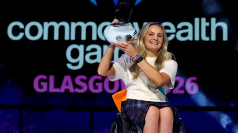 Reuters Melanie Woods holding the David Dixon Award in front of the Commonwealth Games logo. She is smiling broadly.