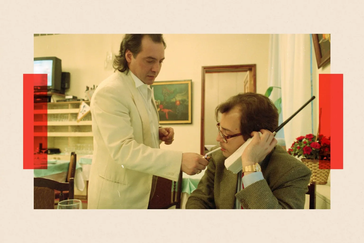 Getty Images A waiter in a white jacket lights the cigarette of a man sitting at a table holding a large old-style mobile phone