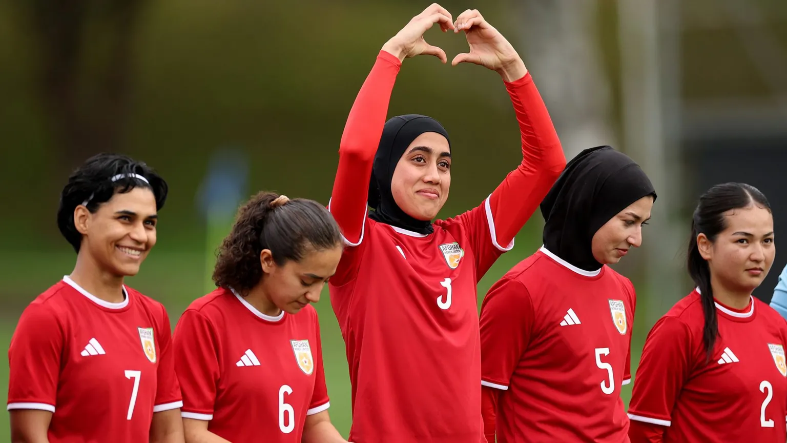 Phil Walter/Getty Images Five women in red football kit stand in a line. The one in the middle is holding her arms above her head and her hands form the shape of a heart.