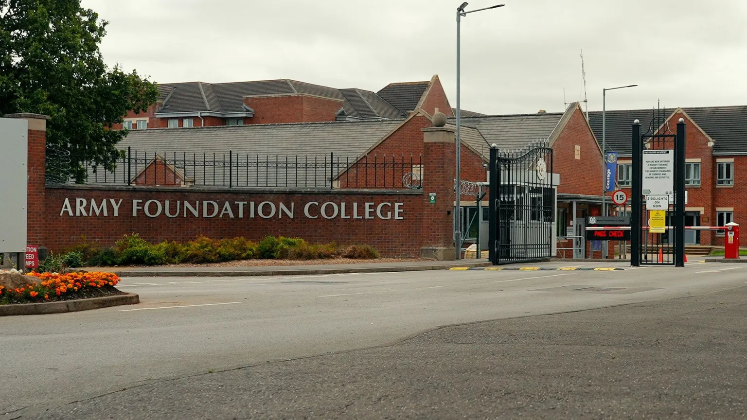 The words Army Foundation College are on a brick wall at the entrance to the campus. Large black gates are open with a red and white barrier slightly beyond.  The college's buildings are behind. 