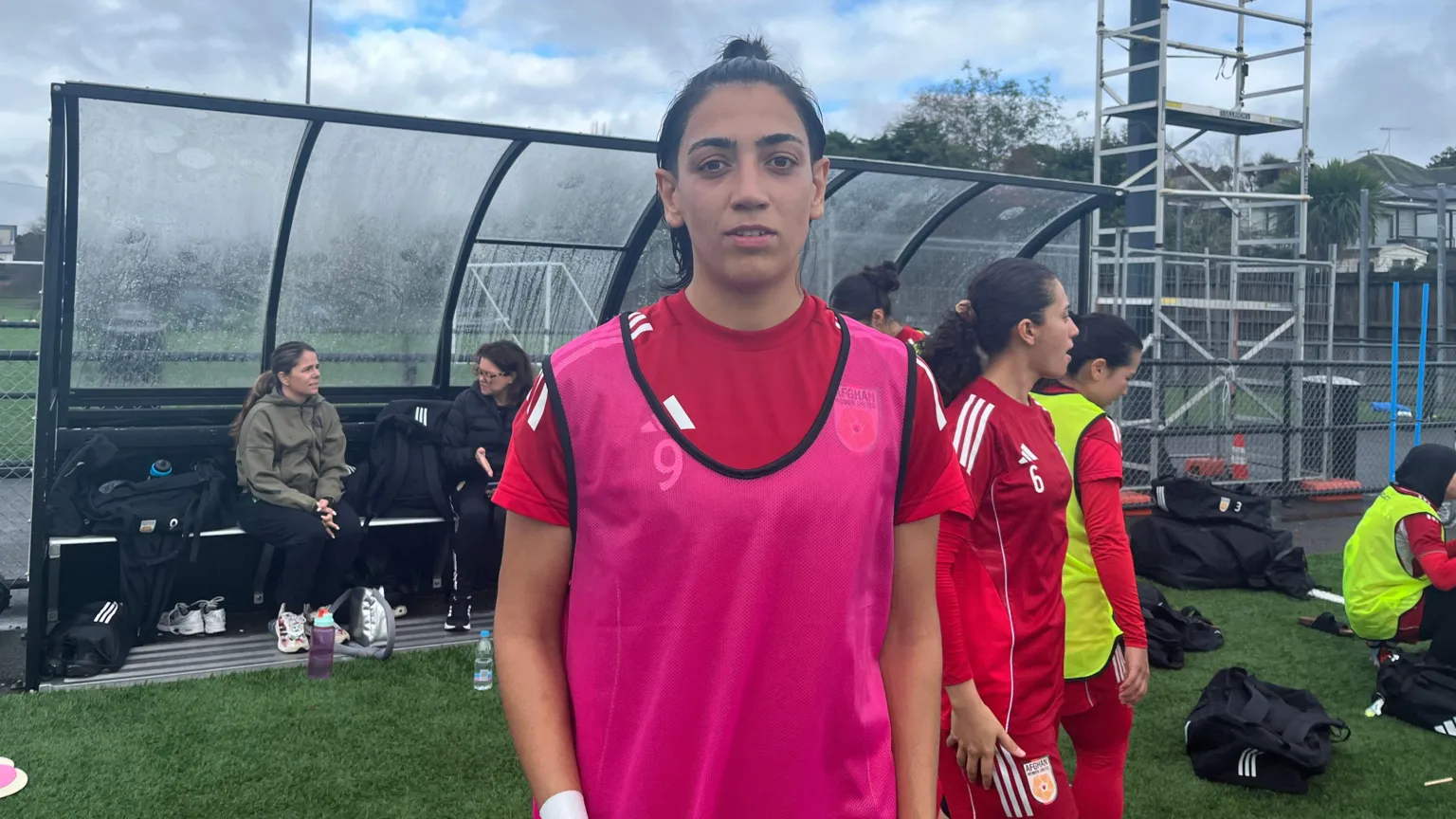 Manozh Noori standing on the edge of a pitch looking at the camera. She has shortish dark hair that is tied back and wears a bright pink vest over her red football kit. In the background there are some kit bags, a few other players and a couple of women sitting in a shelter. The sky is cloudy.