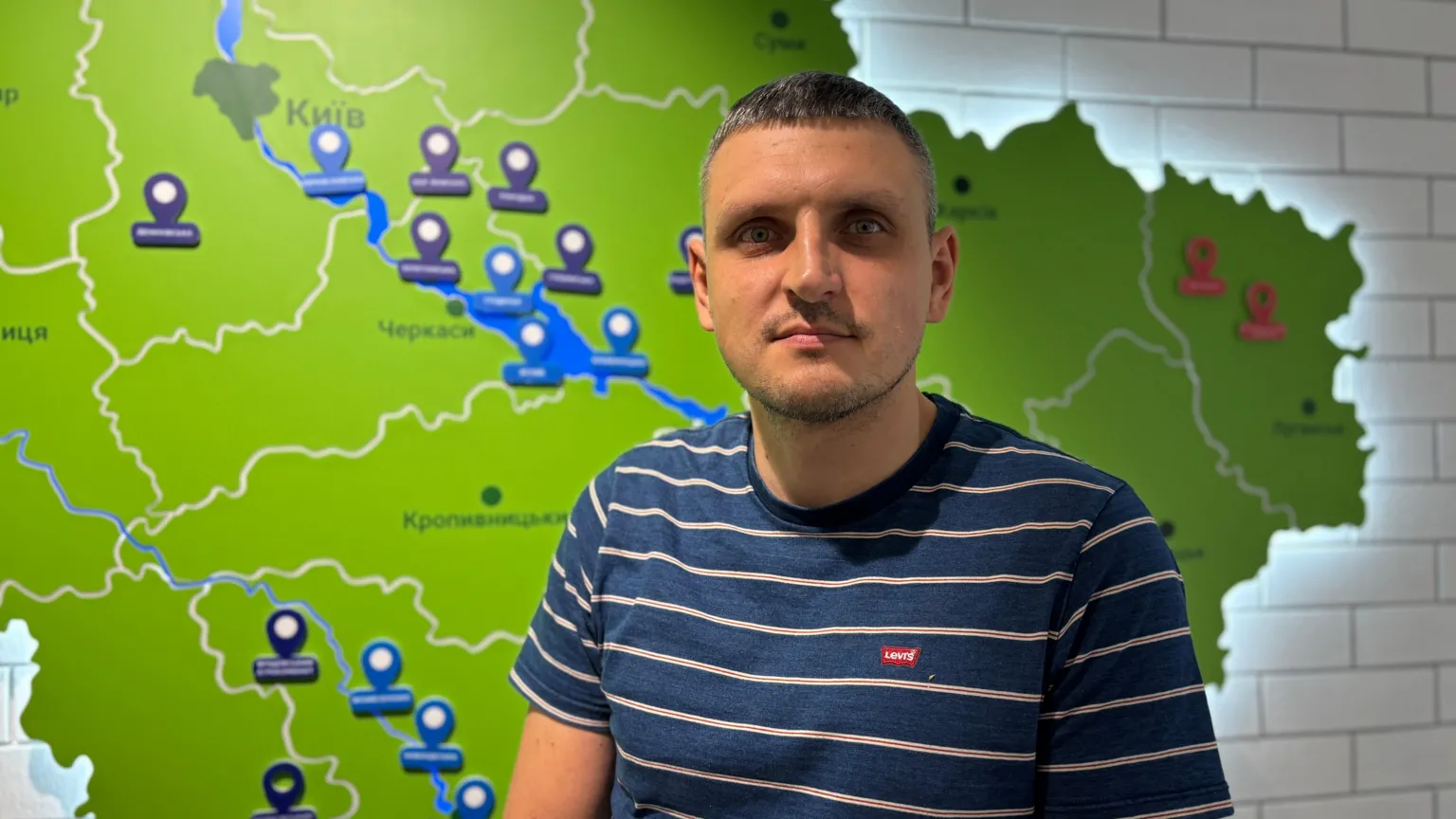 Kristina Volk/BBC A man in a striped blue and white t-shirt stands in front of a map of Ukraine, which is lit up on the wall behind him.